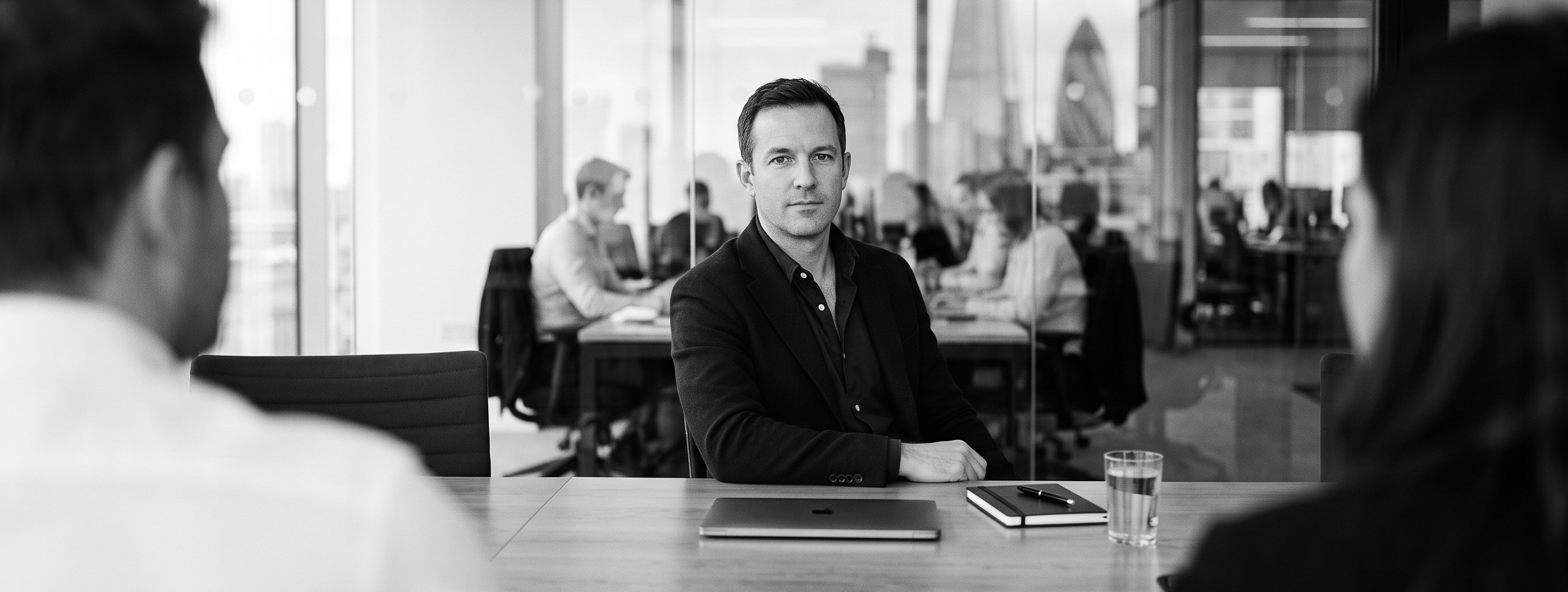 A man at a meeting table in a glass-walled office, colleagues in the foreground